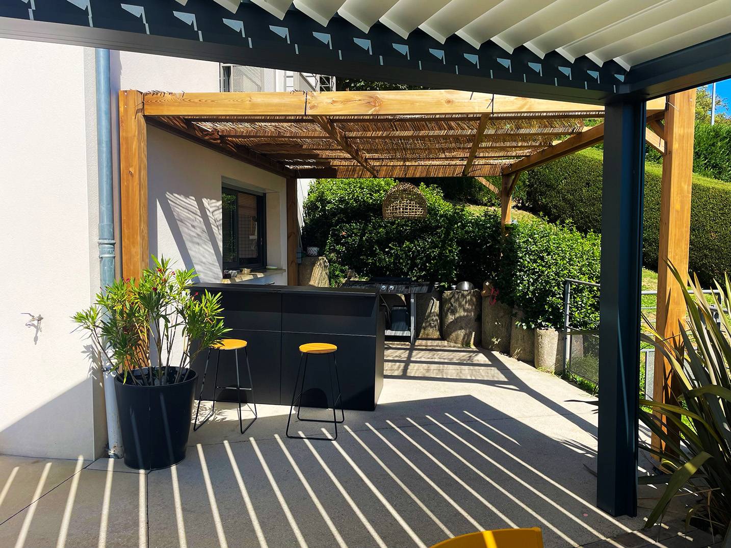 Cuisine extérieure aménagée sous une pergola en bois sur une terrasse attenante à la maison.