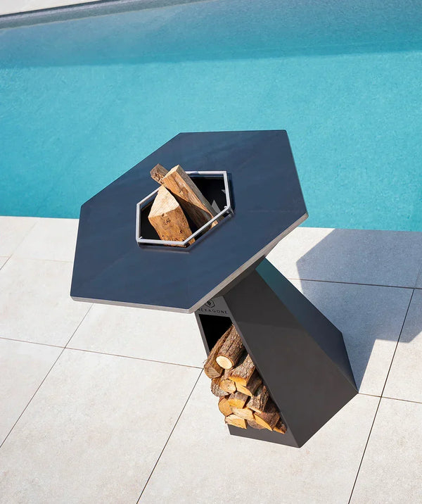 Brasero design hexagonal en acier noir avec rangement à bois, installé en bord de piscine sur une terrasse contemporaine.