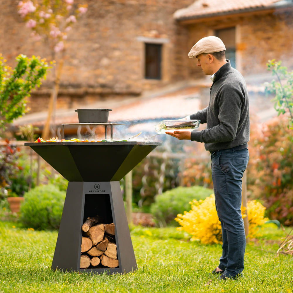 Brasero plancha hexagonal en acier corten avec rangement à bois, utilisé pour la cuisson en extérieur dans un jardin paysager.