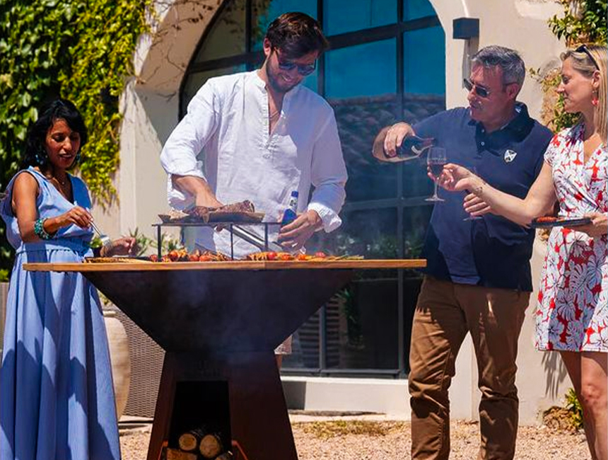 Groupe de personnes cuisinant et partageant un repas autour d’un brasero plancha Hexagone dans un jardin.