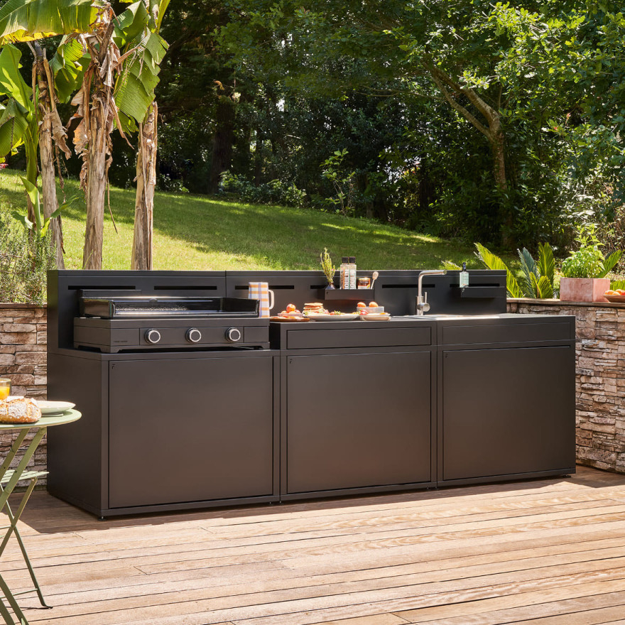 Cuisine extérieure modulaire noire Forge Adour avec plancha, rangements et évier, installée sur une terrasse en bois dans un jardin arboré.
