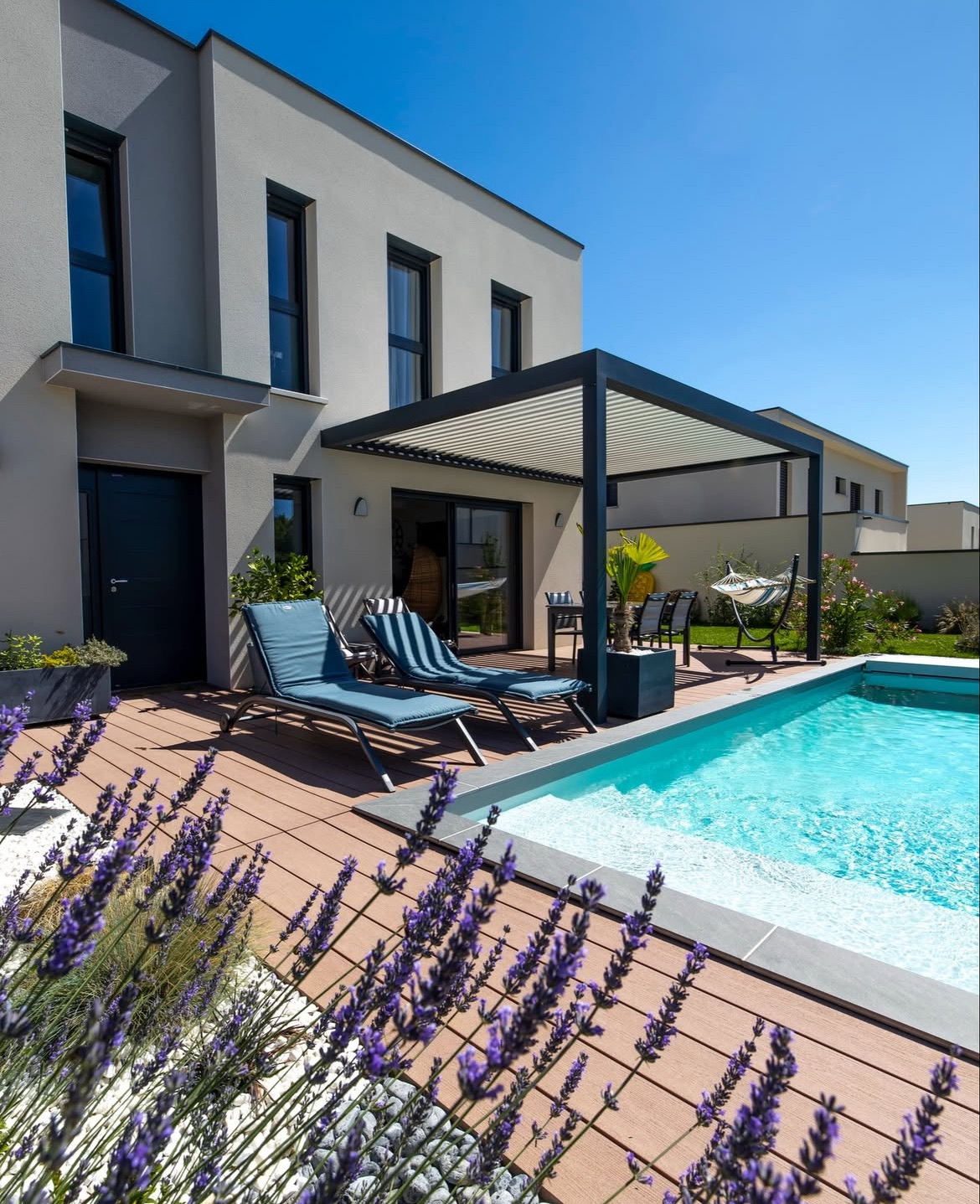 Pergola bioclimatique Galaxie installée sur une terrasse en bois au bord d’une piscine, attenante à une maison contemporaine.